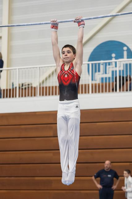Young gymnast in red and black leotard hangs from high bar with focused expression during training session