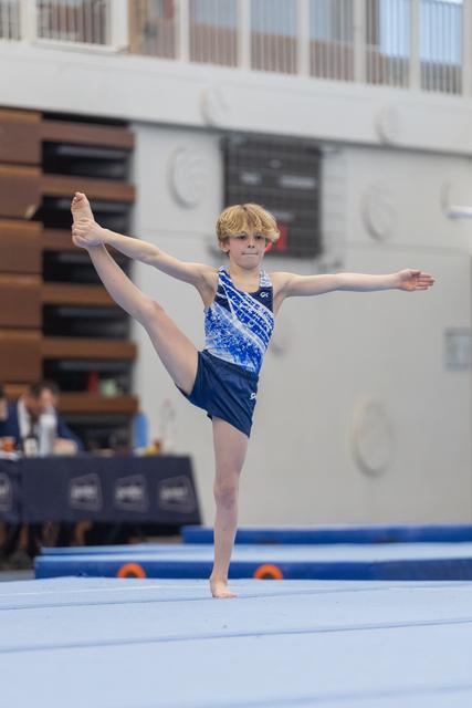 Young male gymnast executes a standing split on floor exercise, demonstrating flexibility and balance in competition setting