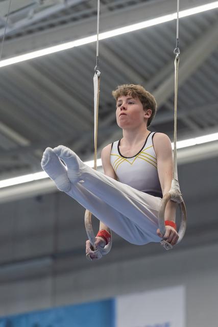 Young gymnast performing on rings apparatus, executing an L-sit hold with focused determination in indoor training facility
