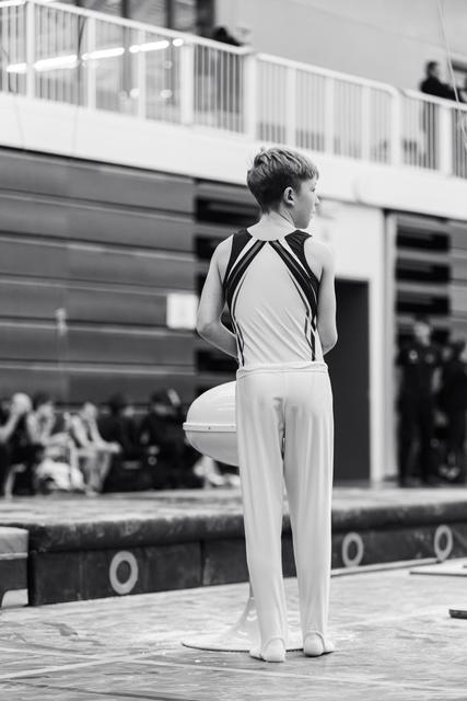 Young male gymnast stands at attention on floor mat, awaiting his turn while spectators watch from bleachers