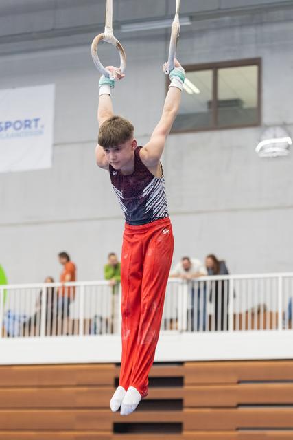 Young gymnast in burgundy and blue leotard with red pants demonstrates strength hold on still rings during routine