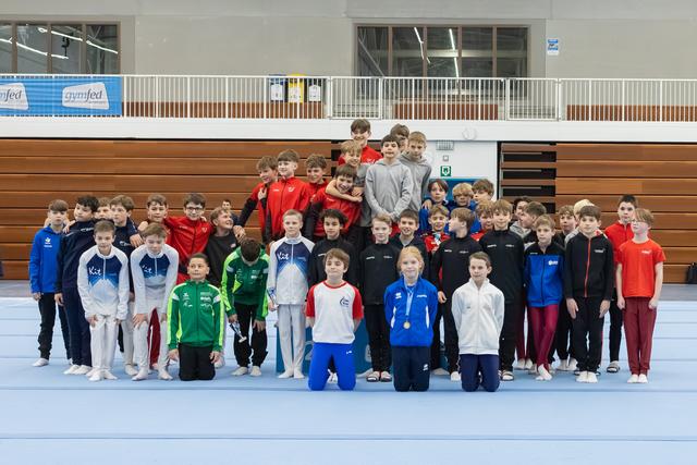 Large group of young gymnasts in colorful tracksuits posing together on blue mat in gymnasium with wooden bleachers