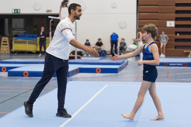 Coach in white shirt greets young gymnast with high-five on training floor, both smiling warmly in gymnasium setting