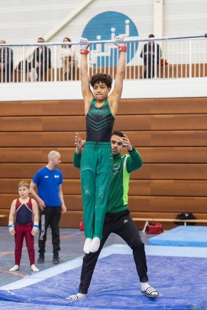 Young gymnast balances upright on coach's shoulders with arms raised, demonstrating acrobatic partnership in gymnasium