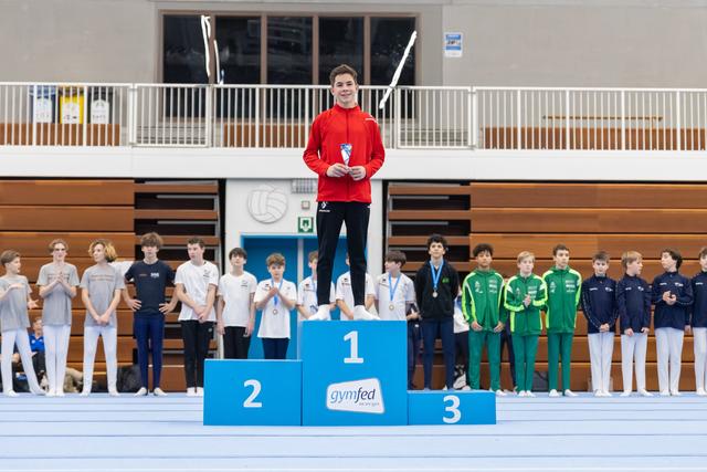Young gymnast in red tracksuit stands atop first place podium holding trophy, with competitors on lower tiers in gymnasium