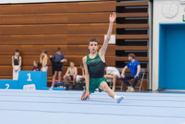 Male gymnast performs lunge pose with arm raised during floor exercise routine in gymnasium