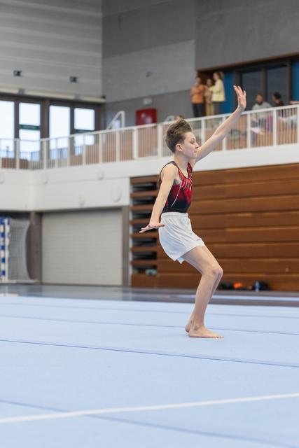 Young gymnast performs floor routine with arms raised and knees bent during landing, wearing red leotard and white shorts