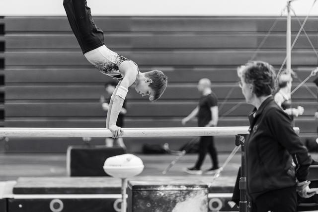 Gymnast performs focused handstand on balance beam while coach observes closely from sideline during practice session