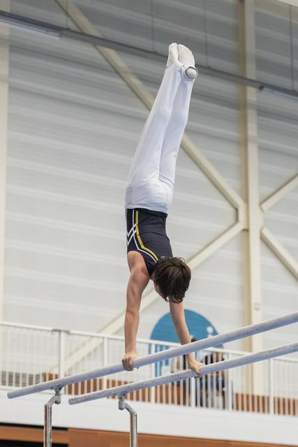 Gymnast performing a vertical handstand on parallel bars, legs extended upward, in an indoor training facility