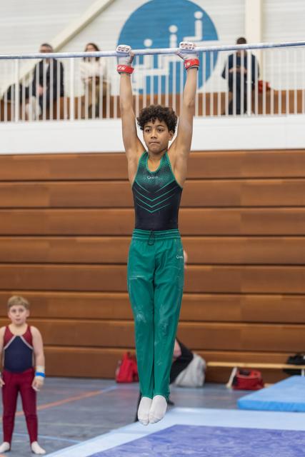 Young gymnast in teal uniform hanging from horizontal bar with focused expression during routine at indoor gym