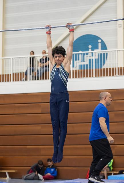 Young gymnast in navy uniform hangs from high bar with coach observing, demonstrating strength in training facility