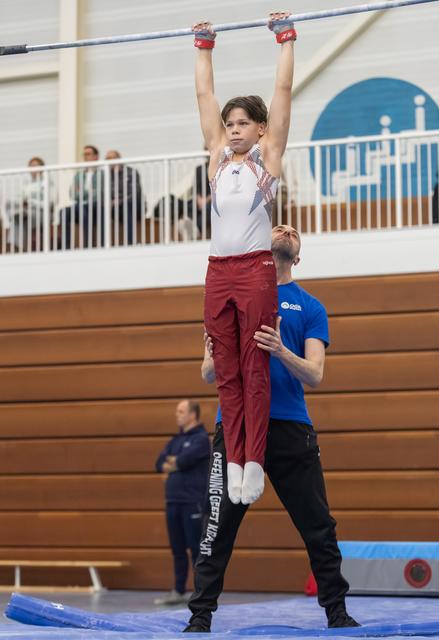 Young gymnast hangs from high bar with focused expression while spotter stands ready below in indoor training facility