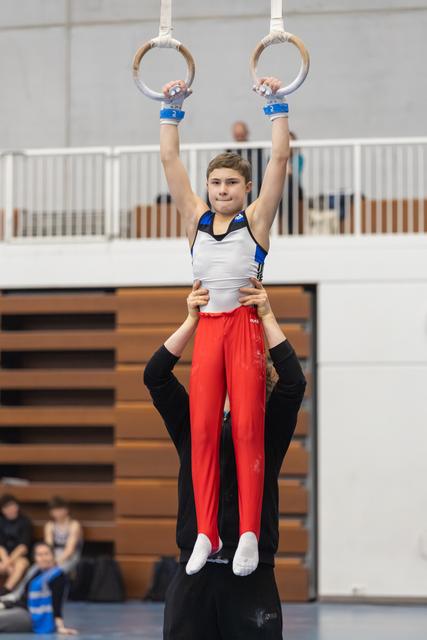 Young gymnast in red pants hangs from rings while coach supports from below in gymnasium