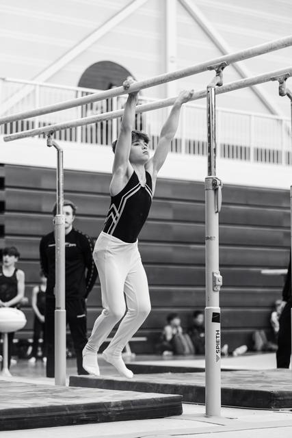 Young gymnast in black and white attire hanging from uneven bars with focused determination during training in gymnasium