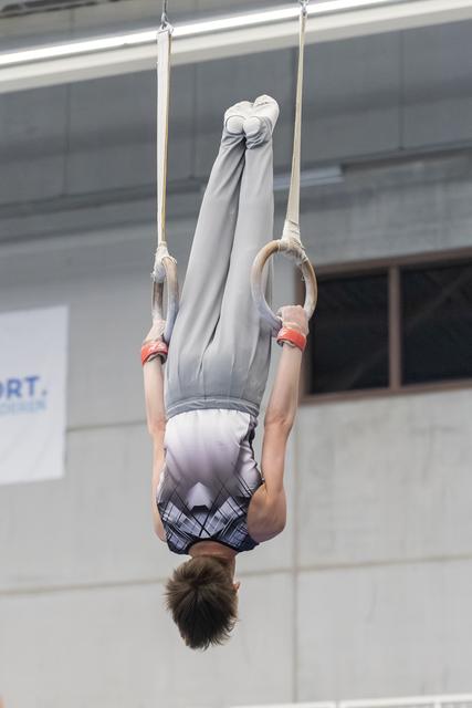 Young gymnast performing an inverted hang position on still rings, displaying strength and control in a gray competition uniform