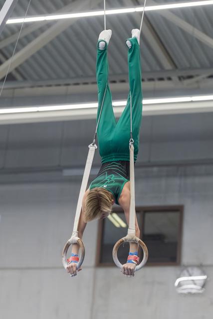 Young gymnast performs vertical hang on rings, displaying strength and control while wearing teal uniform in indoor facility