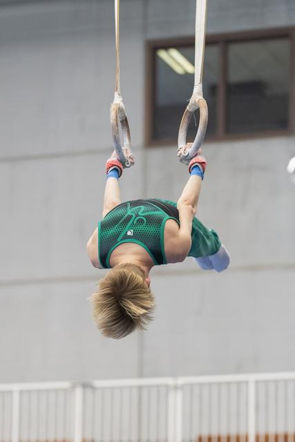 Young gymnast performing inverted hang on still rings, body arched backward in green leotard during apparatus routine