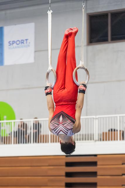 Gymnast performs inverted hang on rings with extended body, wearing red outfit during indoor training session