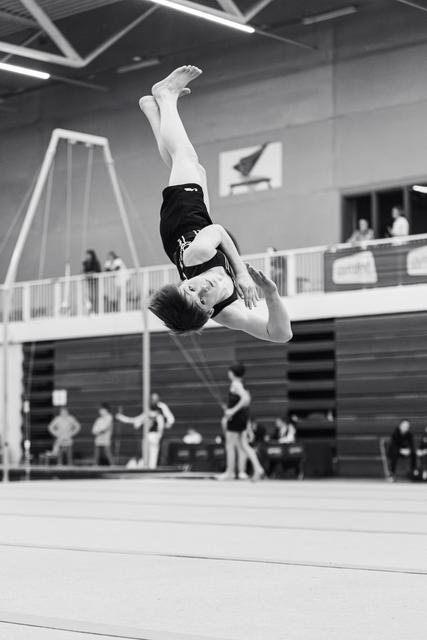 Gymnast executing a tucked backflip mid-air on the floor exercise mat in an indoor training facility