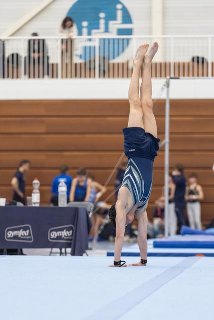 Gymnast executing a precise handstand during floor routine, legs extended vertically, in gymnasium with spectators and officials