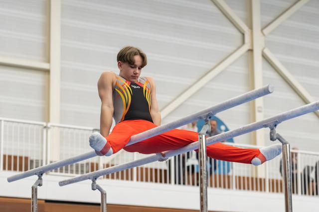 Male gymnast executing a horizontal hold on parallel bars, wearing orange and black leotard with focused expression