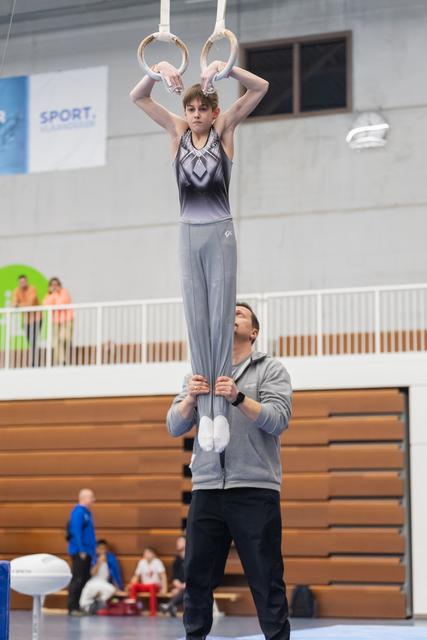 Young gymnast holds rings position with coach supporting from below in an indoor training facility