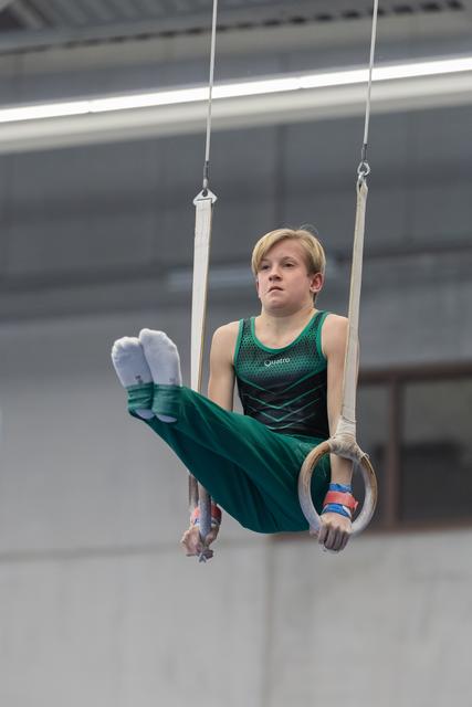 Young gymnast in green leotard performing L-sit position on rings, displaying intense focus and strength during routine