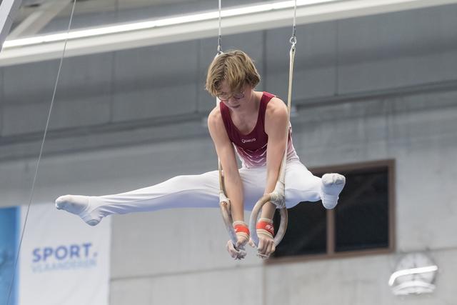 Young gymnast executing a wide straddle hold on rings with intense concentration, wearing maroon leotard in indoor training facility