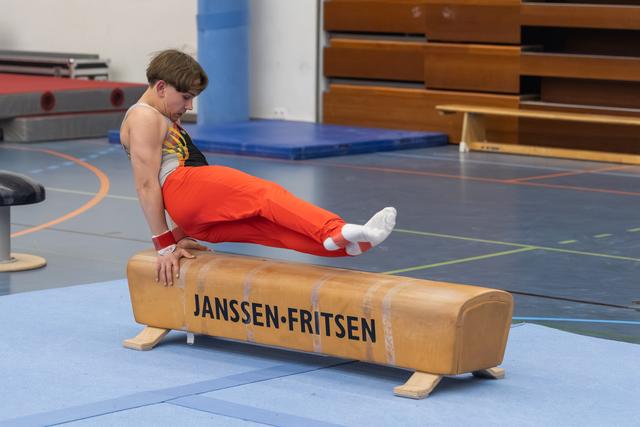 Male gymnast in orange pants executes a skill on the pommel horse, extending his legs horizontally during practice