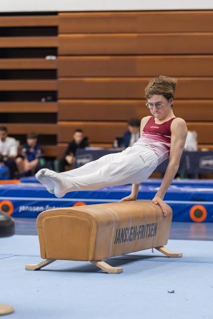 Male gymnast performs L-position hold on pommel horse during routine, demonstrating strength and control in white and maroon uniform