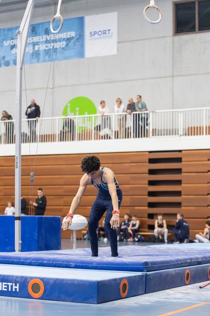 Male gymnast in navy leotard prepares for dismount from rings, hands gripping apparatus in indoor gymnasium