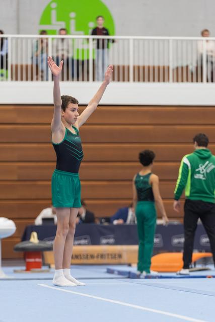 Gymnast in green and navy leotard stands with arms raised overhead, completing a floor routine at an indoor gymnastics meet.