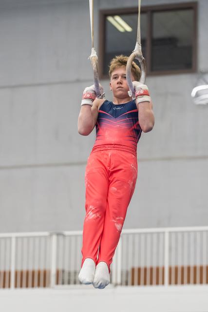 Young gymnast in coral outfit performing still rings routine, displaying intense focus and strength during apparatus work
