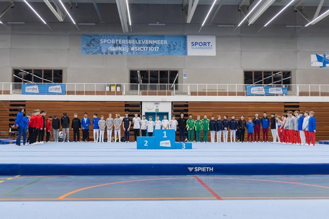 Multiple gymnastics teams lined up on award podium and floor exercise area during medal ceremony in sports hall