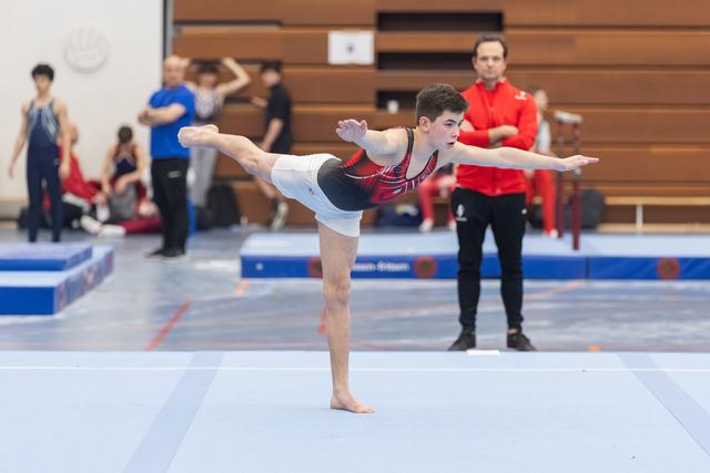 Male gymnast performs an arabesque balance on floor exercise, arms extended gracefully, while coaches observe from the sidelines