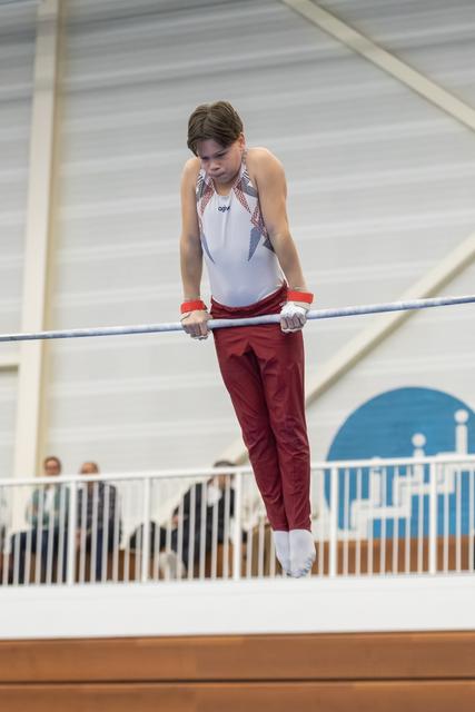 Male gymnast in white and maroon uniform performs on horizontal bar with focused expression, spectators visible in background
