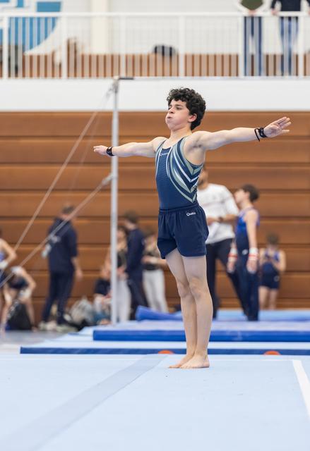 Male gymnast performs floor routine with arms outstretched in blue and white leotard at indoor gymnasium