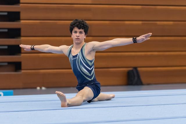 Male gymnast performs floor routine in split position with arms extended, wearing teal leotard in indoor training facility