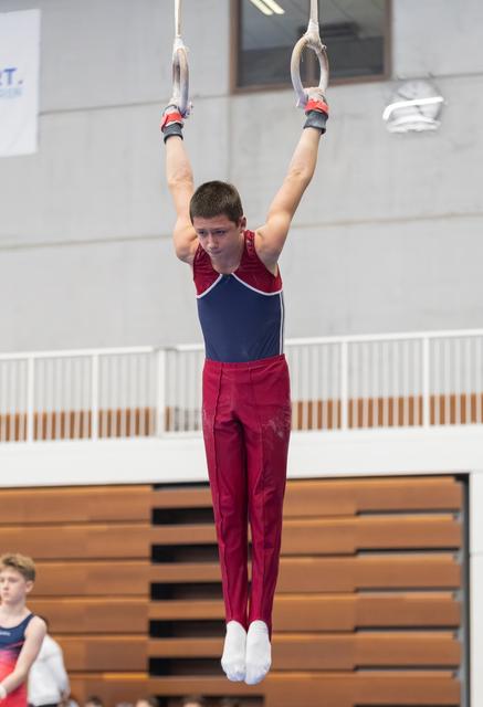 Male gymnast in maroon and navy uniform performs a strength hold on rings, arms extended overhead in gymnasium