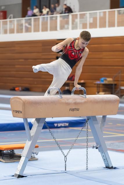 Male gymnast performs pommel horse routine with extended leg, displaying strength and control in red and black leotard