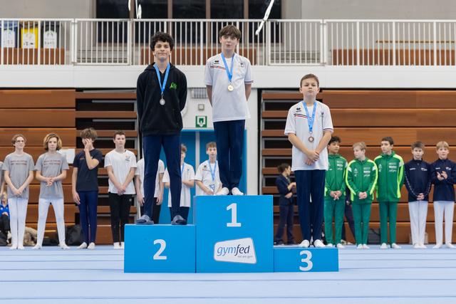 Three young gymnasts stand on award podium wearing medals, with first place center, teammates watching from background