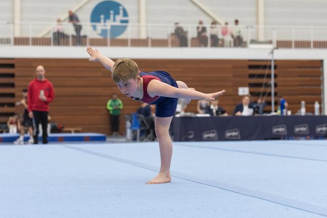 Young gymnast performs arabesque balance on floor exercise, arms extended, focused expression at indoor gymnastics meet