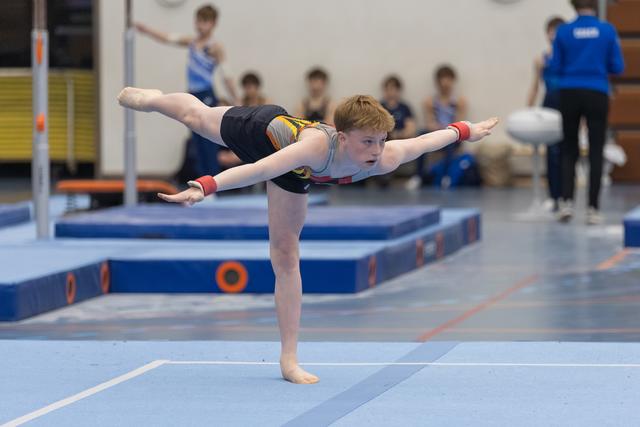 Young gymnast in black leotard performs arabesque balance on floor, arms extended with focused expression during routine