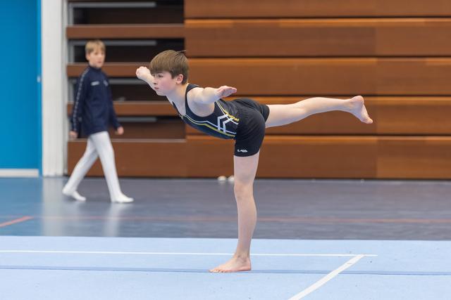 Young gymnast holds an arabesque balance on the floor, arms and leg extended, with another athlete walking in background