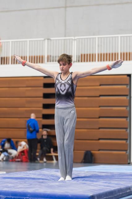 Young male gymnast stands with arms extended horizontally on floor mat, displaying control and form during routine