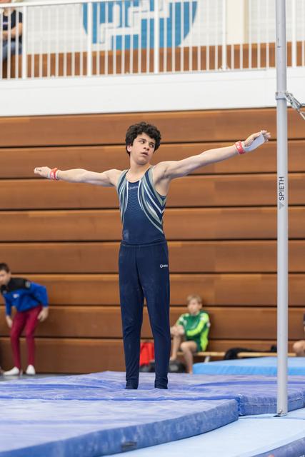 Young gymnast in navy leotard stands with arms outstretched in preparation pose on mat, focused expression before routine