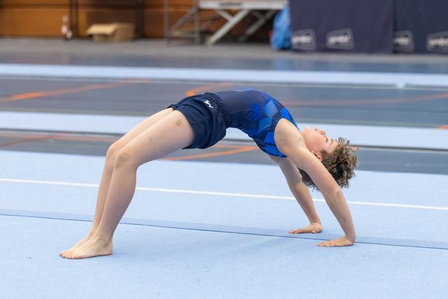 Young gymnast in blue leotard performs a backbend bridge on training floor, demonstrating flexibility and strength