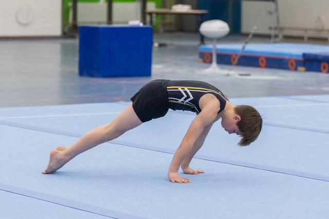 Young gymnast in black and gold leotard performs a bridge stretch on blue floor mat during training session
