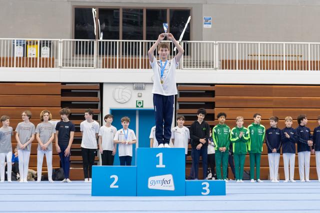 Young gymnast raises trophy overhead on first place podium while teammates stand below in gymnasium