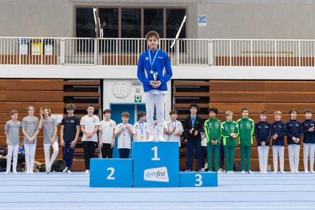 Young gymnast in blue tracksuit stands atop first place podium holding medal, with competing teams lined up behind